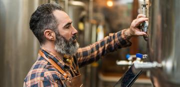 Image shows a man with a beard looking at a dial on a brewing tank. He appears to be checking measurements in a craft brewing warehouse, surrounded by metal containers